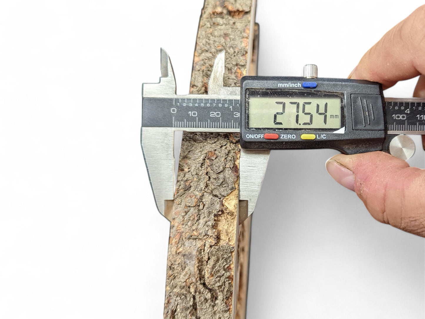 Digital caliper held by a hand measures the rough bark edge of a wood slice, showing 27.54 mm.