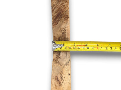 A yellow tape measure gauges the width of a narrow, rustic timber with mottled grain.