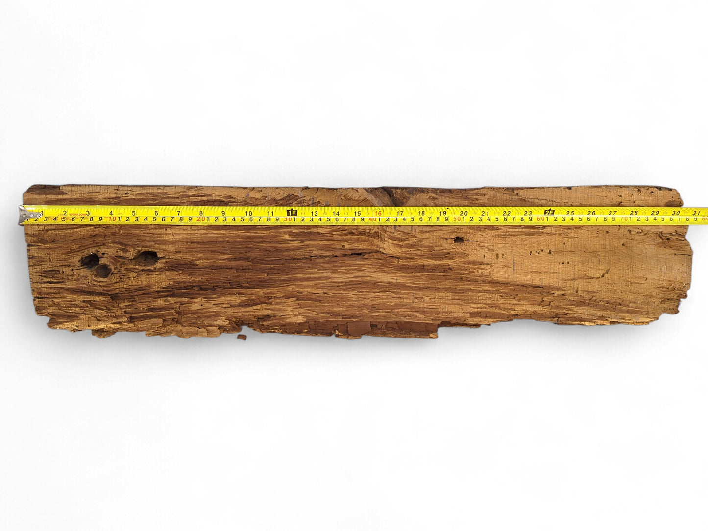 Rough, dark brown reclaimed wood beam fragment with flaking, crumbly texture and a yellow measuring tape.