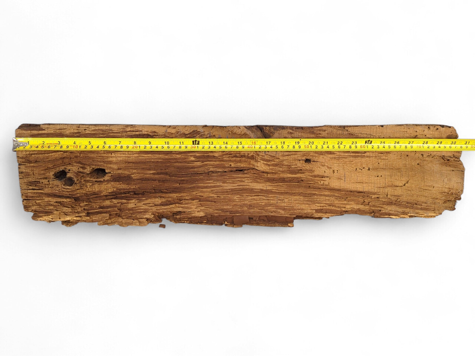 Rough, dark brown reclaimed wood beam fragment with flaking, crumbly texture and a yellow measuring tape.
