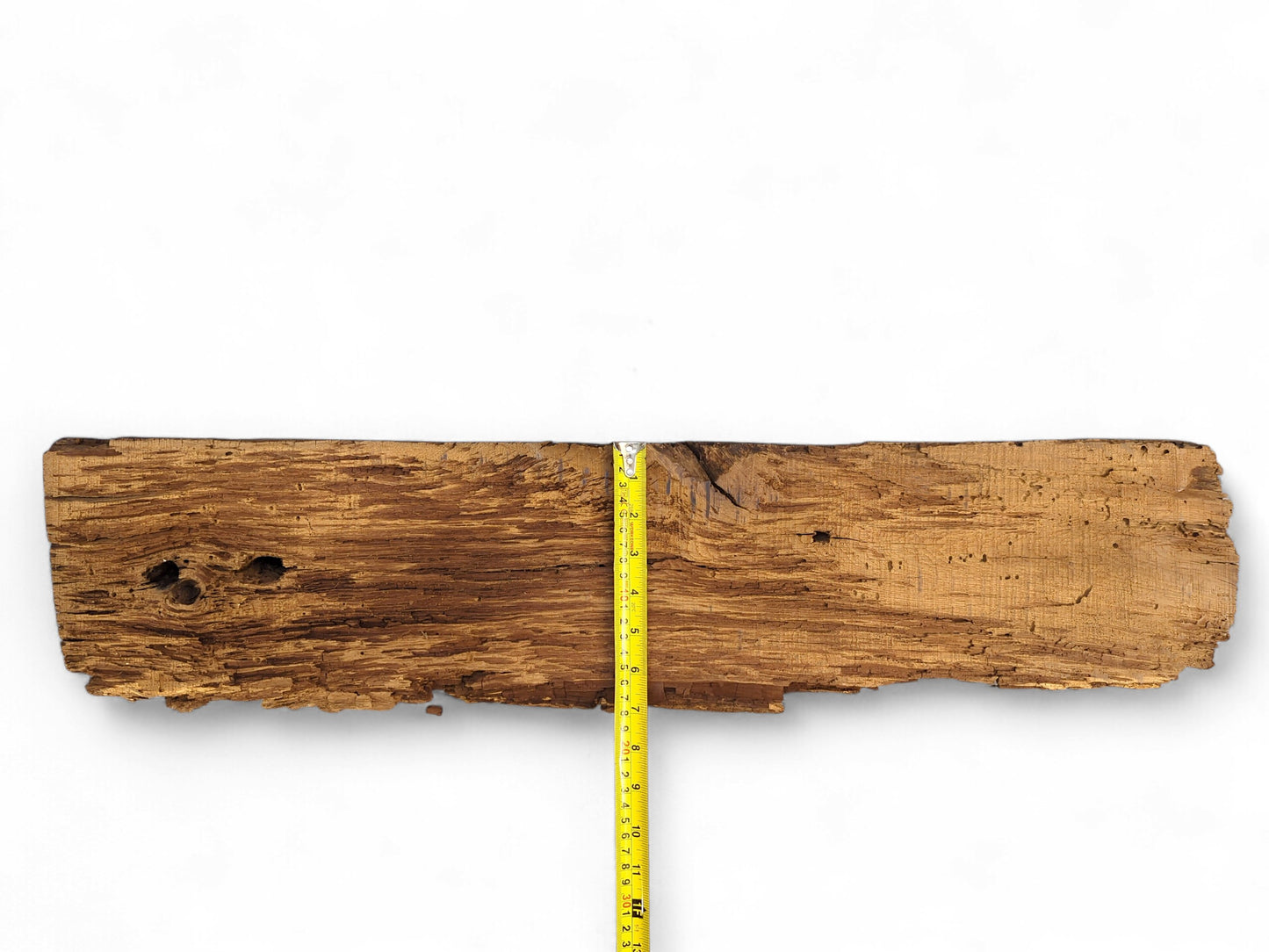 Rough, aged brown wood fragment features deep cracks, a flaking surface, holes, and a yellow measuring tape.