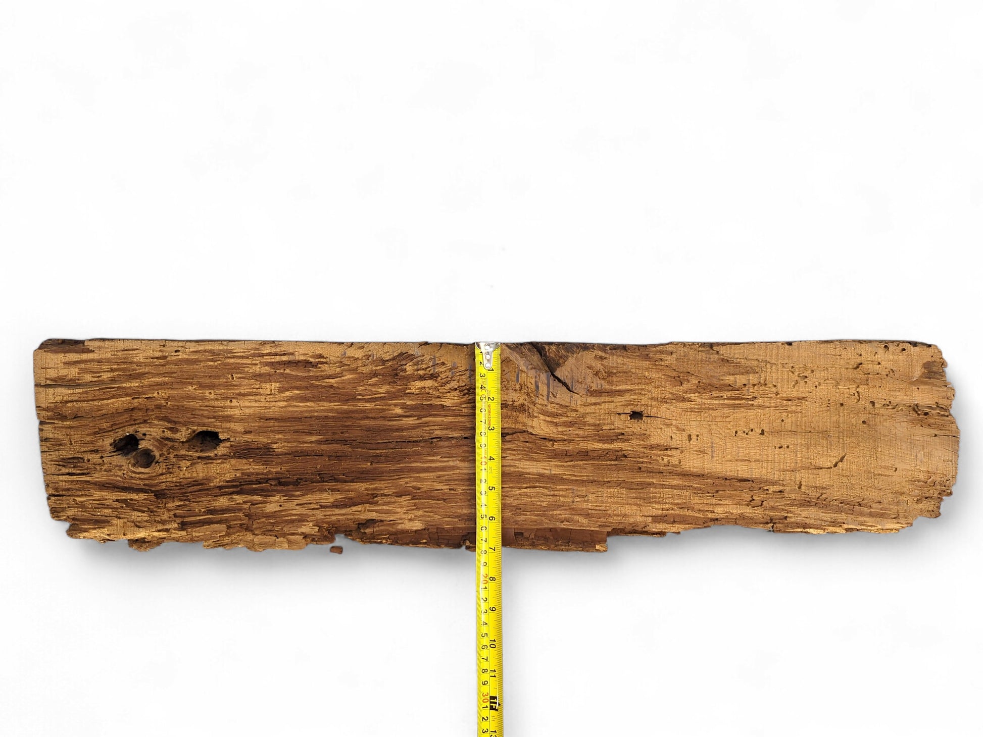 Rough, aged brown wood fragment features deep cracks, a flaking surface, holes, and a yellow measuring tape.