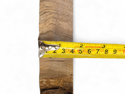 Rough-textured wood slice with light brown tones and wavy grain, horizontally measured by a yellow tape.