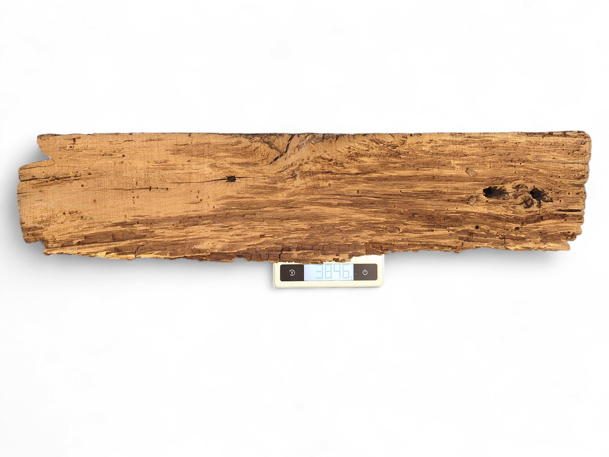 Weathered brown wood fragment showing extensive cracks, boreholes, and a flaking surface, resting on a digital scale.