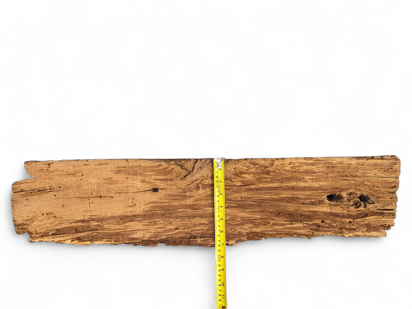 Textured brown wood fragment displaying deep fissures, extensive flaking, boreholes, and a vertical yellow tape measure.