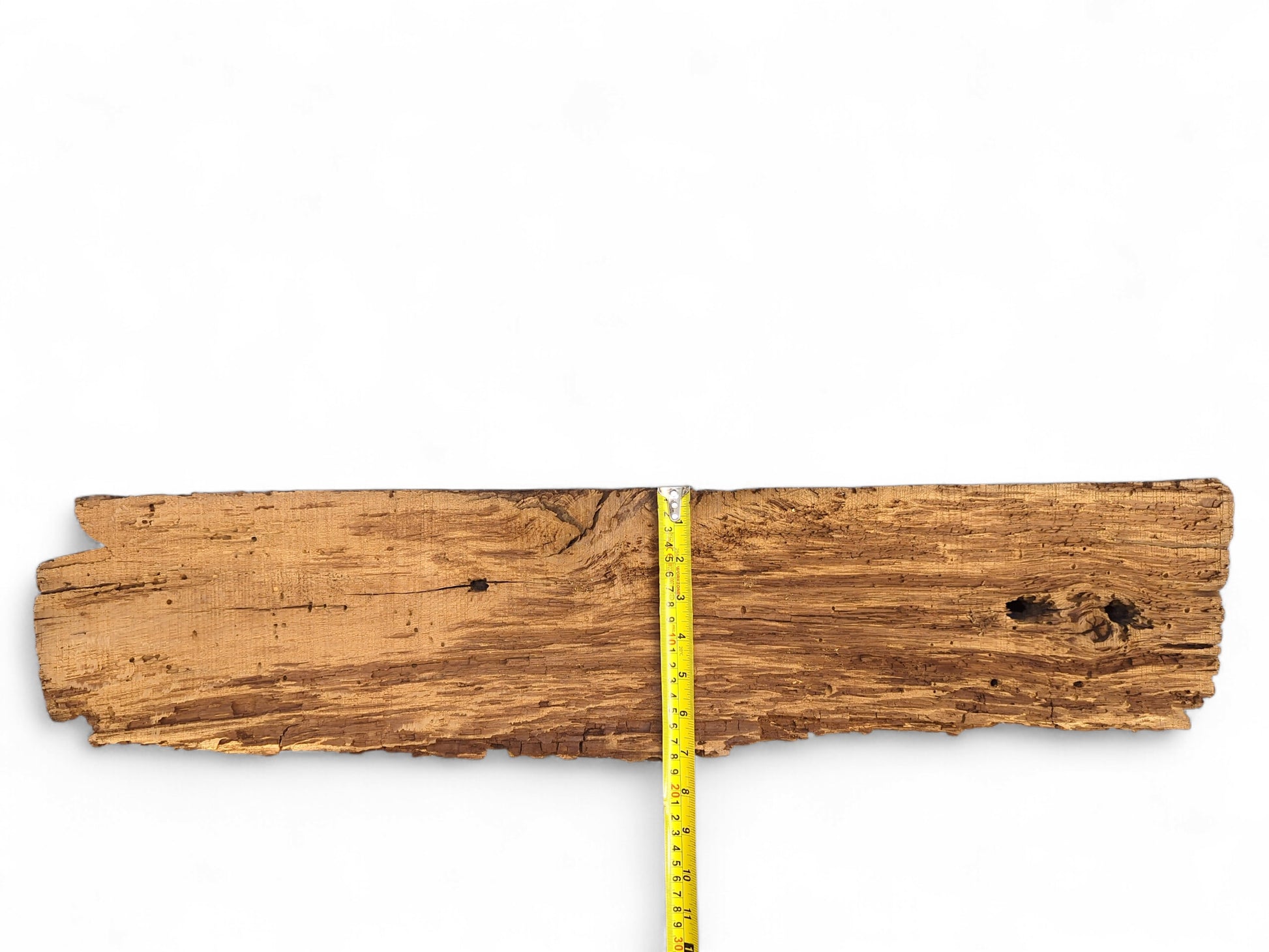 Textured brown wood fragment displaying deep fissures, extensive flaking, boreholes, and a vertical yellow tape measure.