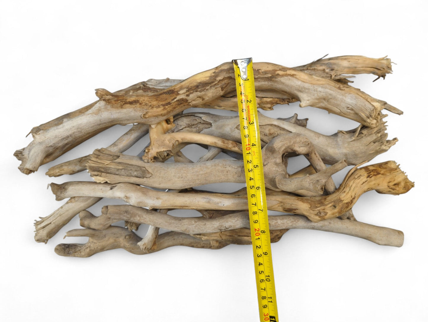Irregular, textured, weathered natural driftwood sticks, light brown and gray, piled, measured by a yellow tape.