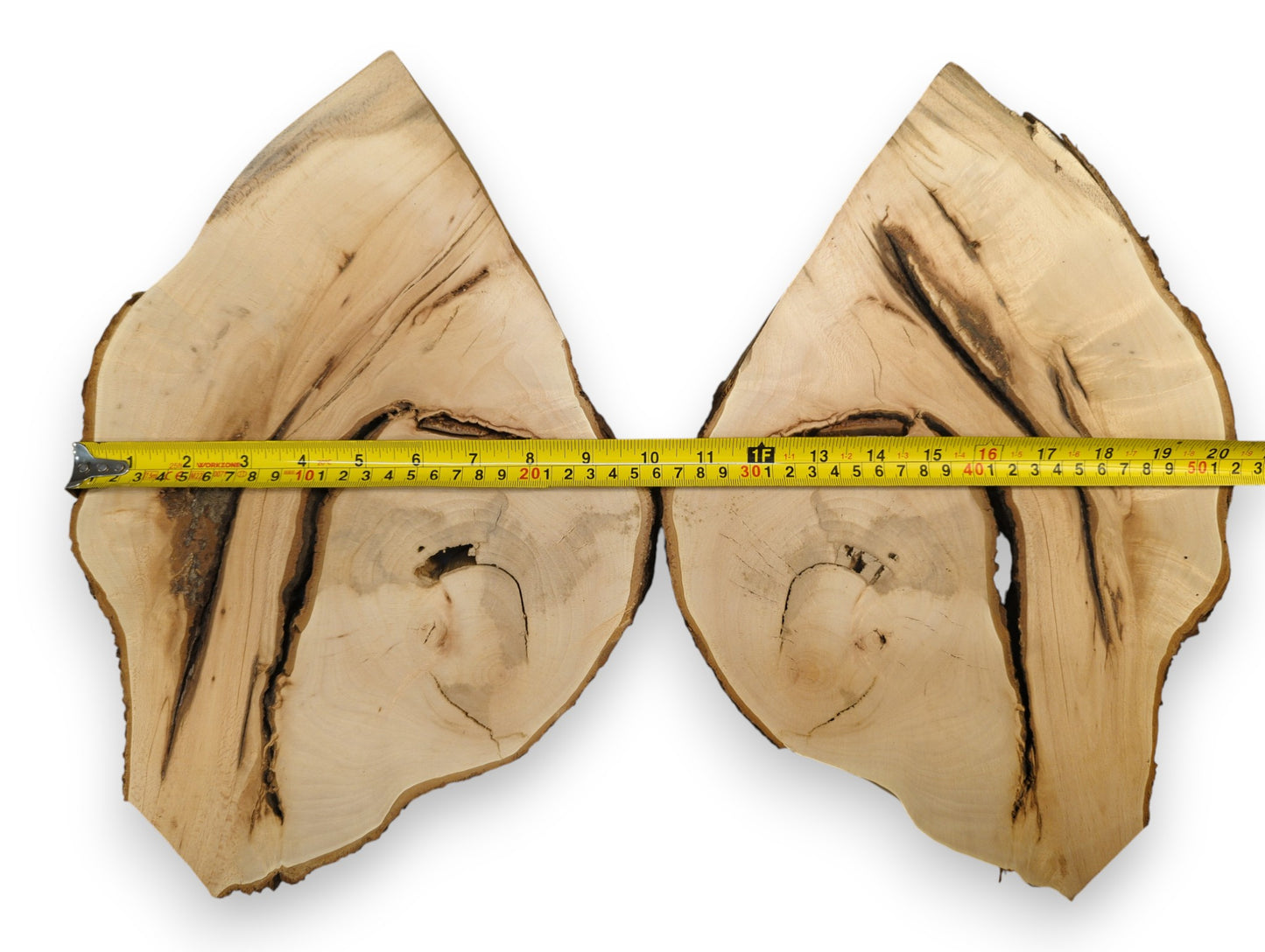 Light-colored bookmatched wood slices display dark irregular grain, natural voids, bark, and a yellow measuring tape.
