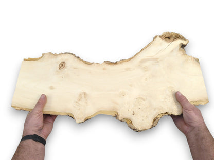 A long, pale wood plank with a rugged, undulating live edge is held by two hands.