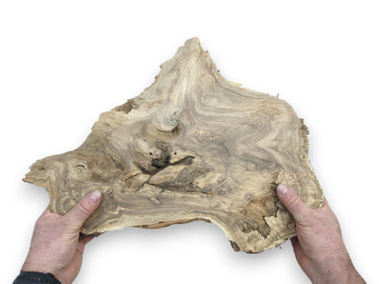 Two hands hold a wide, raw-edged timber slab with marbled grain and a gnarled center.