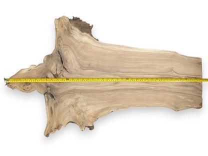 A yellow tape measure spans an irregular timber plank with flowing grain and gnarled edges.