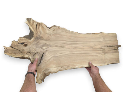 Hands hold a wide timber plank with a gnarled burl and flowing, contoured grain.
