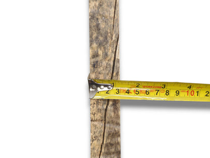 A measuring tape gauges the width of a mottled lumber plank with a deep fissure.