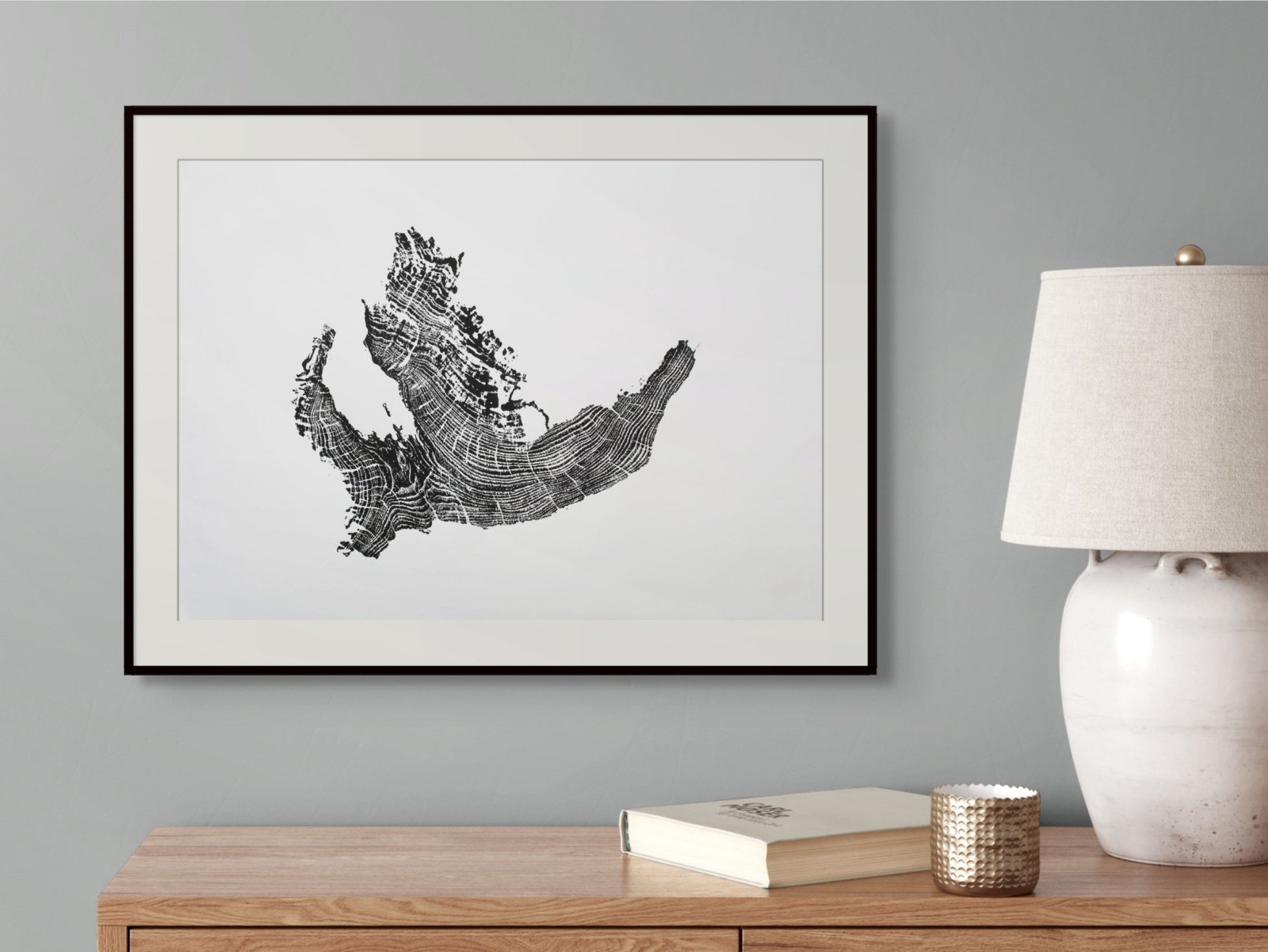 Black framed print of a tree root monoprint with concentric rings, hanging above a wooden side table.