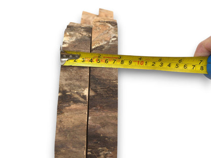 A person measures the horizontal width of two rustic, dark-barked wood planks placed side-by-side.
