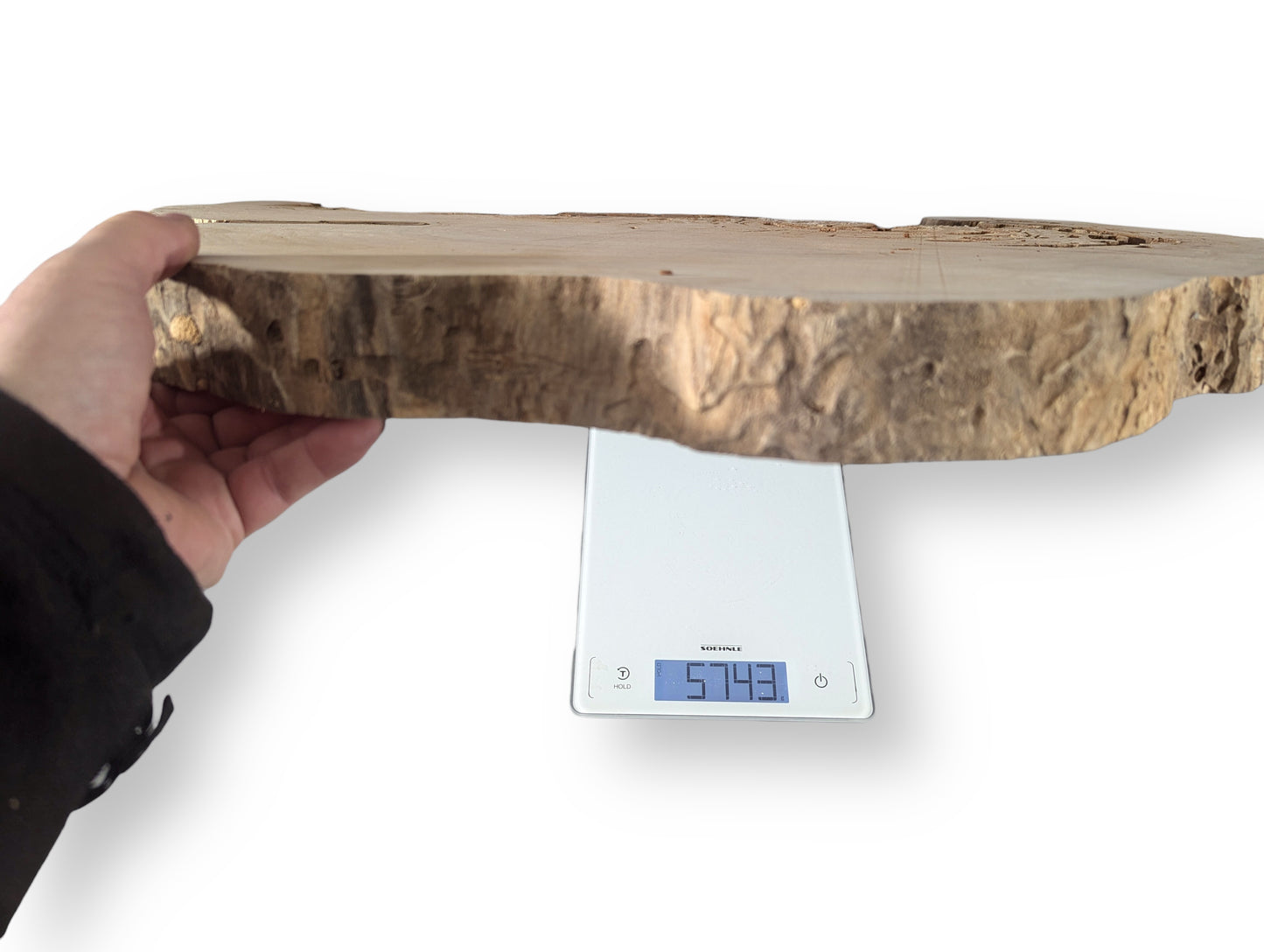 Hand holds an irregular light brown wood slice with rough bark over a digital scale showing 5743.