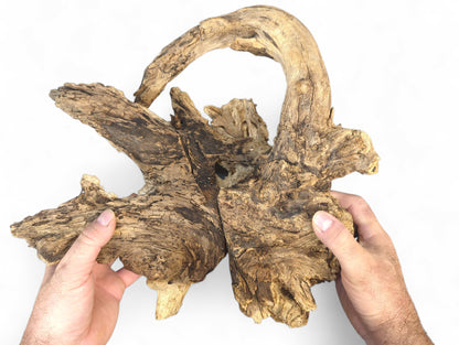 Hands grasp an aged, multi-toned brown wood piece, featuring an arching top and rough, cracked surface.