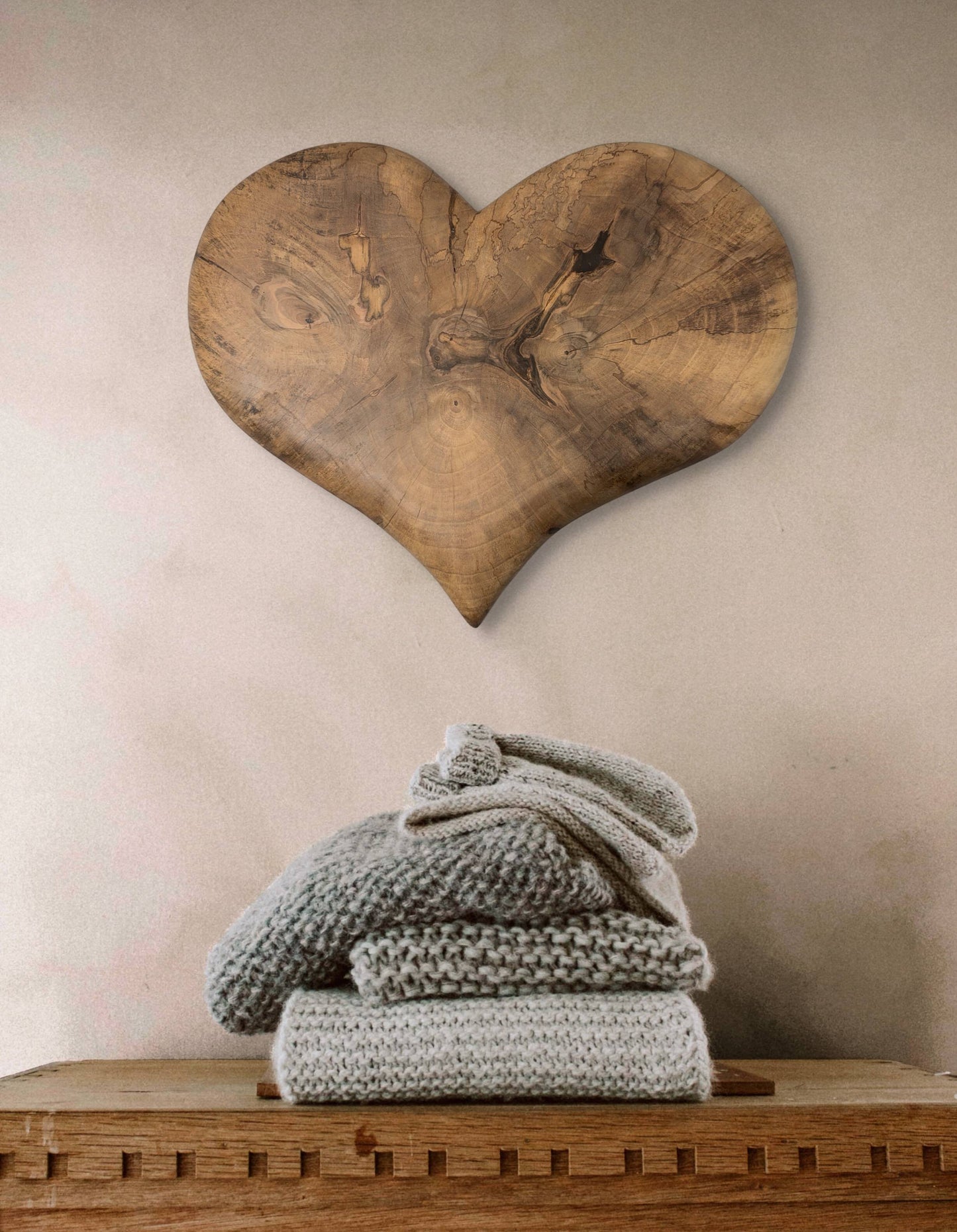 Heart-shaped wooden art piece with natural grain hangs above stacked grey knitwear on a wooden surface.