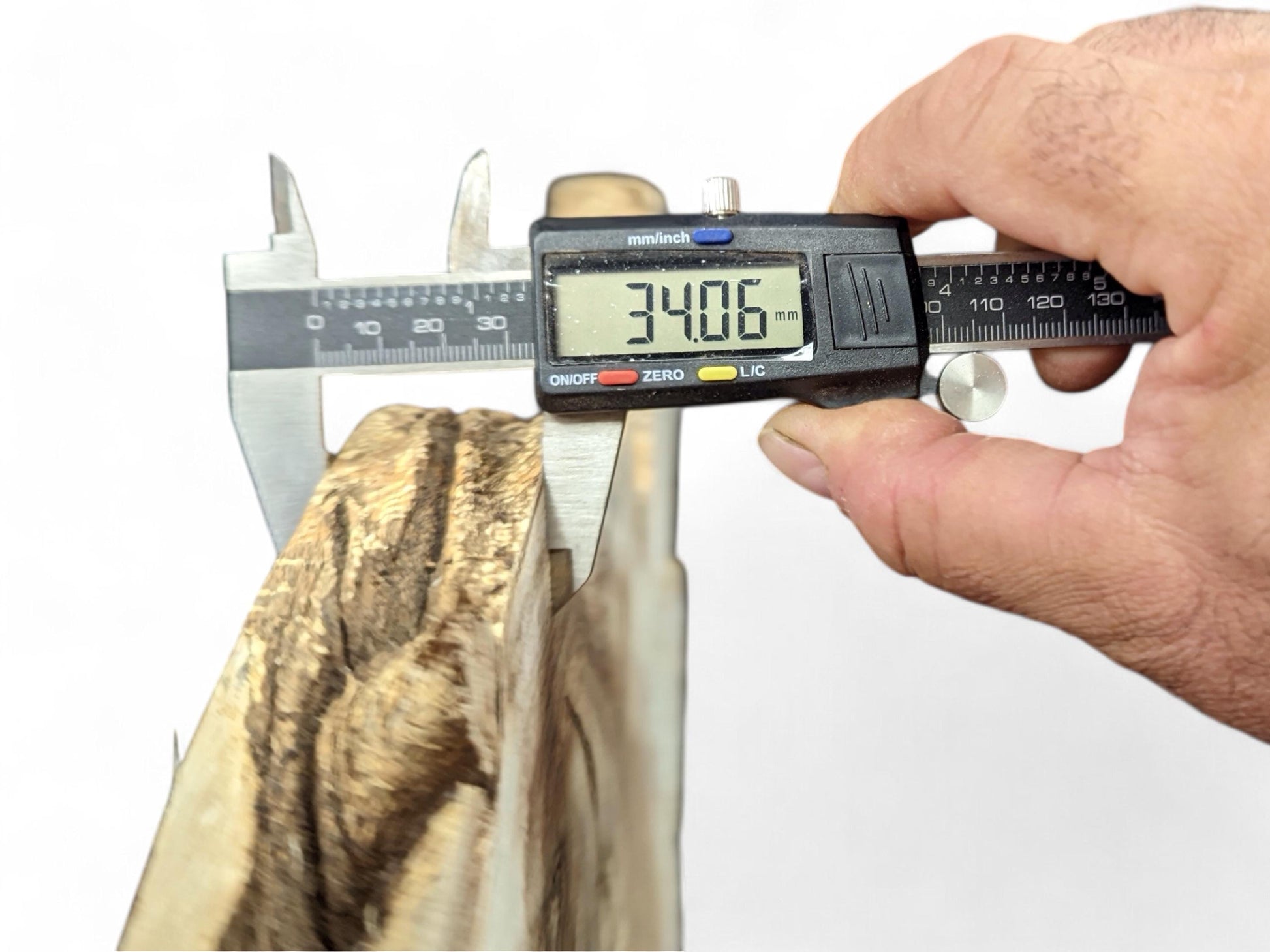 Digital caliper held by a hand measuring the 34.06 mm thickness of an irregular light brown wood slice.