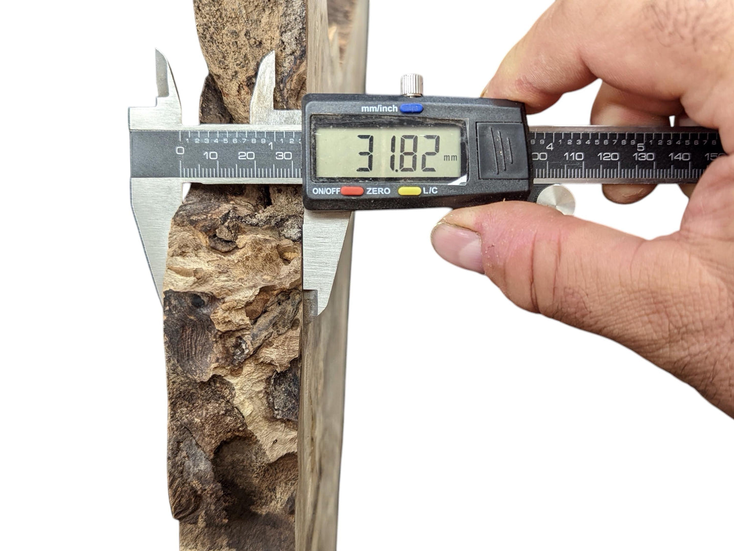 A hand holds a digital caliper, measuring the 31.82 mm thickness of a textured, light brown wood slice.