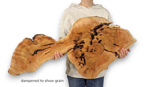 Irregularly shaped, golden-brown live edge wood slab, held by a person. Text: 'dampened to show grain'.