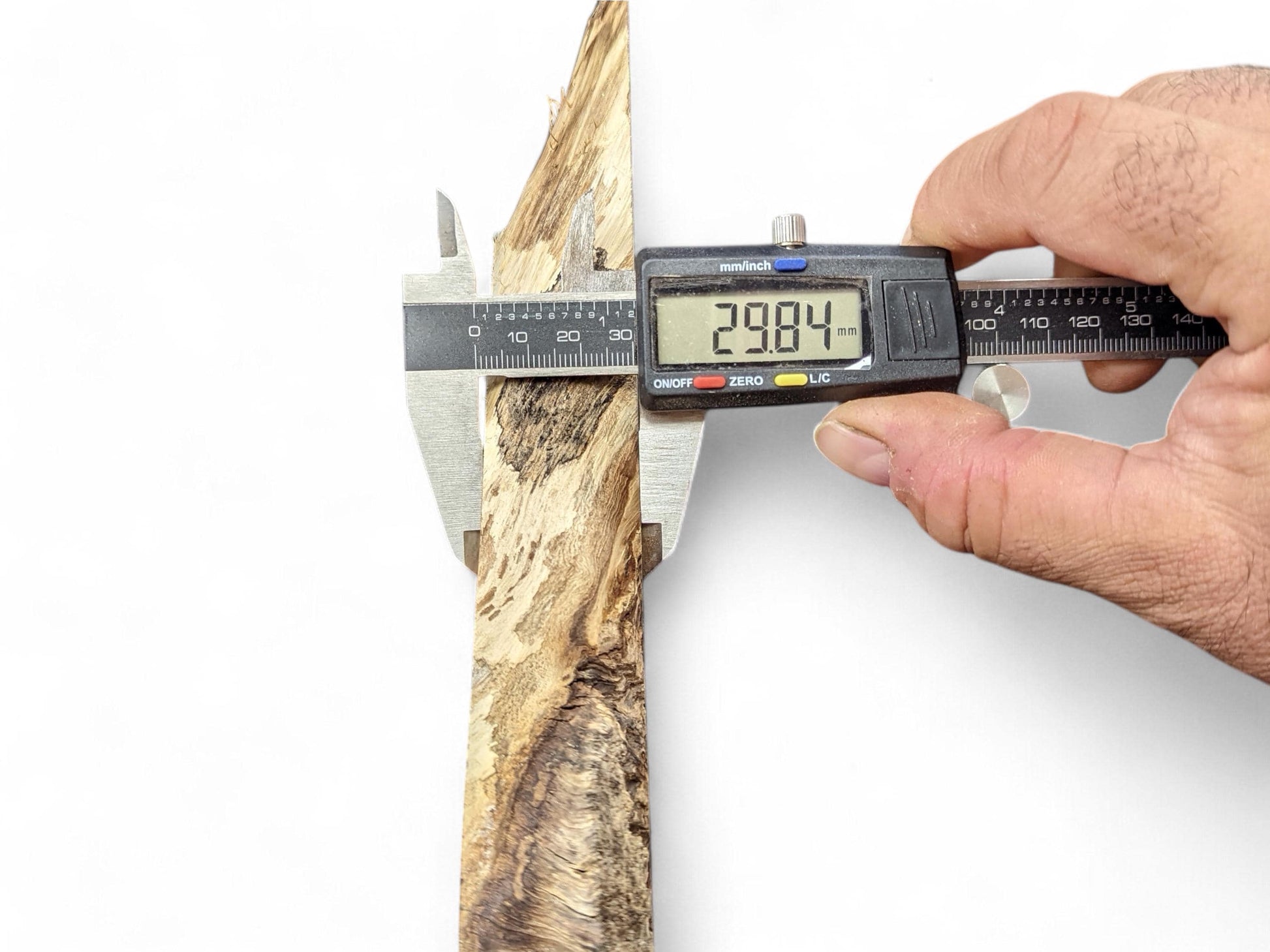 A hand holds a digital caliper, measuring an irregular wood slice displaying 29.84 mm.