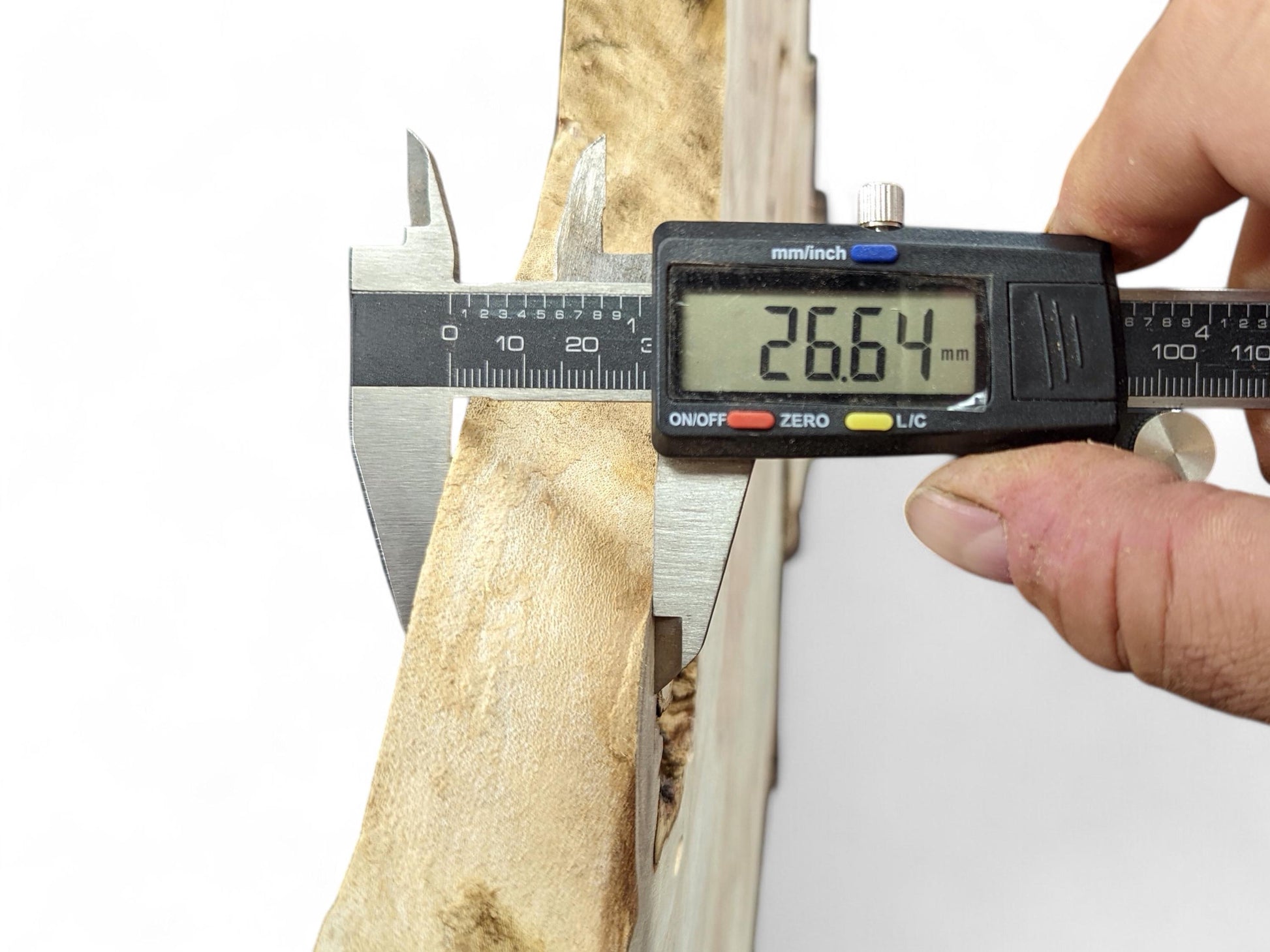A hand holds a digital caliper measuring a light brown wood slice, showing 26.64 mm.