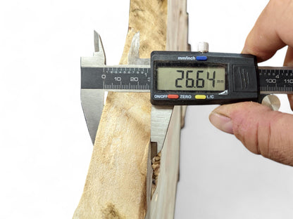 A hand holds a digital caliper measuring a light brown wood slice, showing 26.64 mm.