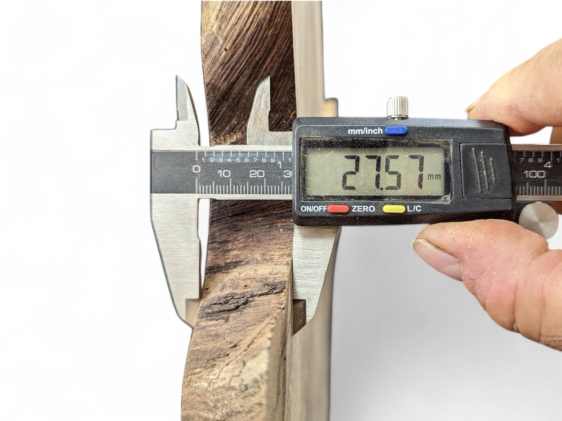 A digital caliper measures a dark brown, rough-edged wood slice, displaying a thickness of 27.57 mm.