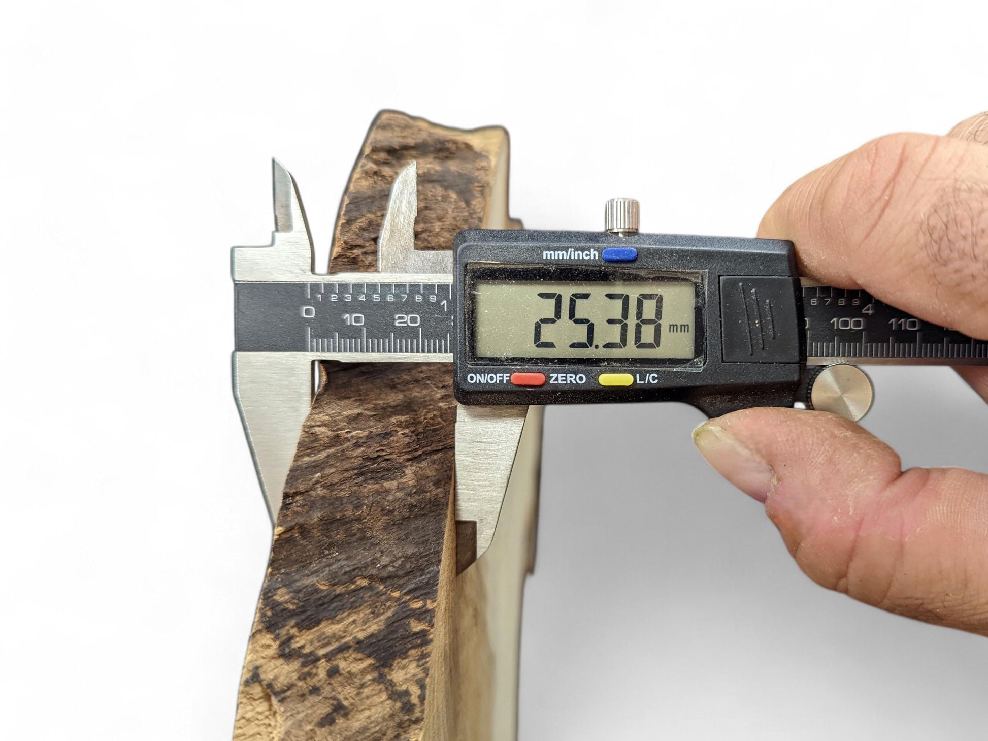 Digital caliper measures a dark brown, irregularly textured wood slice, displaying 25.38 mm thickness.