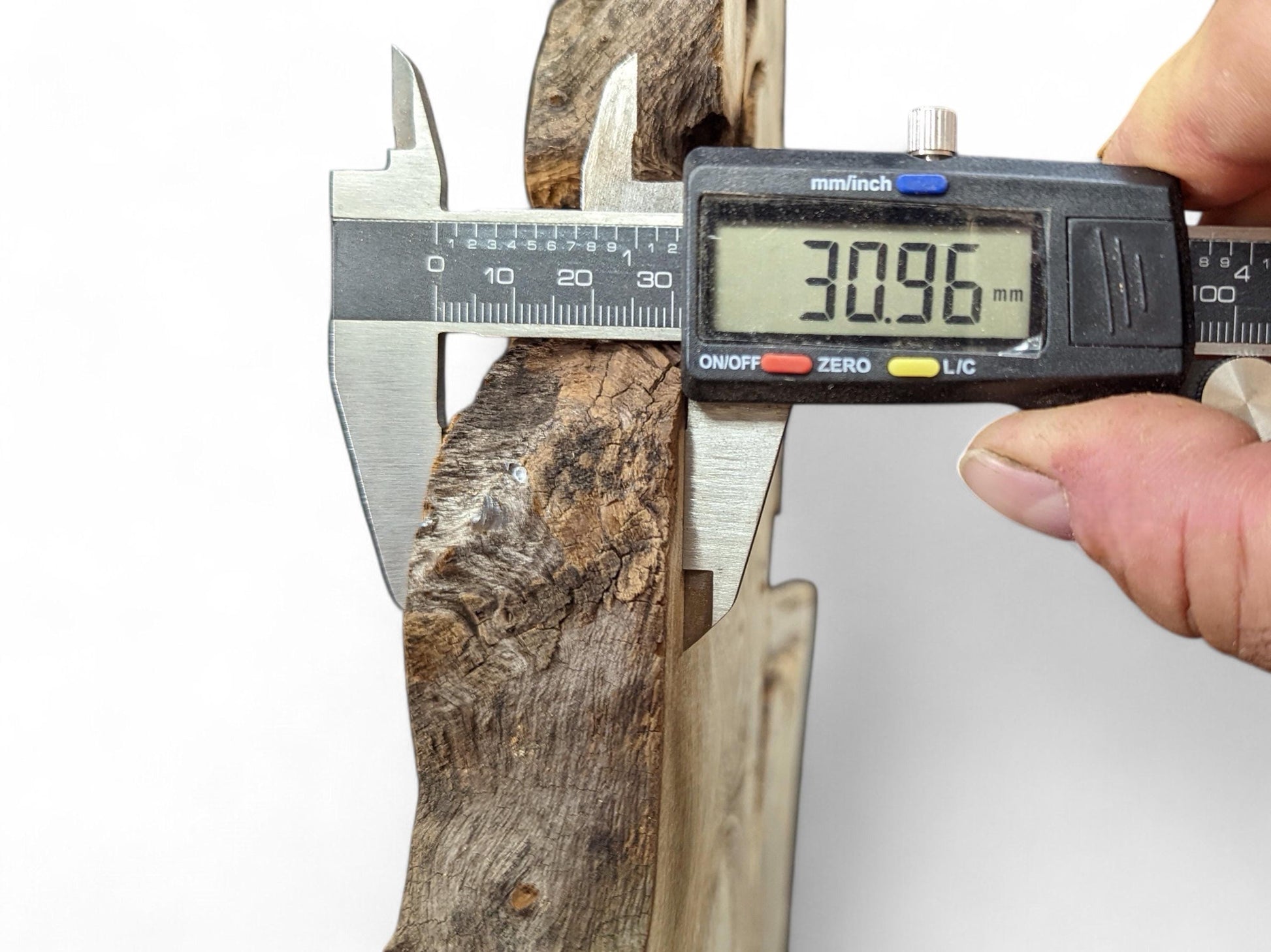 Hand holding a digital caliper, measuring 30.96mm thickness of a dark, textured wood piece.