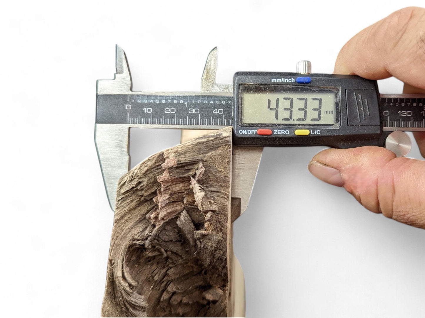 A hand holds a digital caliper, measuring the 43.33 mm thickness of a textured brown wood piece.